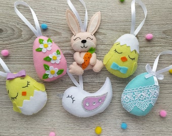 May include: Six felt Easter decorations hanging from white ribbon. The decorations include a pink egg with white flowers, a blue egg with white lace, a yellow chick in an eggshell, a white bird with a pink and silver wing, a yellow chick with a blue bow, and a brown bunny holding a carrot.