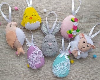 May include: A selection of Easter felt decorations. The ornaments include a yellow chick, a pink bird, a grey bunny with glitter ears, a pink egg with flowers, a white sheep, and two eggs with lace details. Each has a white ribbon for hanging.