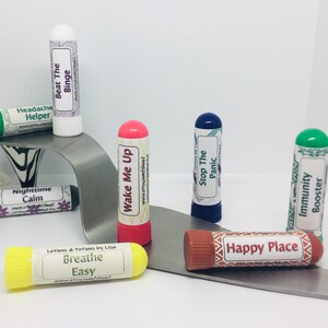 May include: An assortment of lip balm tubes with various colored caps and labels. The labels feature text such as "Headache Helper," "Beat The Binge," and "Happy Place." The products are arranged on a silver, curved surface.
