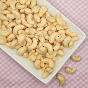 Könnte beinhalten: Eine weiße Keramik Schale gefüllt mit Cashewnüssen. Die Nüsse sind in einem zufälligen Muster angeordnet und haben eine hellbeige Farbe.