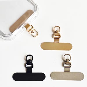 May include: A collection of phone charm holders in gold, black, and silver. Each holder features a rectangular base with a loop for attaching a charm or strap. One is attached to a clear phone case.