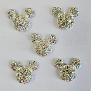 May include: Five Mickey Mouse head-shaped embellishments covered in iridescent rhinestones. The rhinestones reflect a spectrum of colours. The embellishments have a silver base and are arranged on a light grey surface.