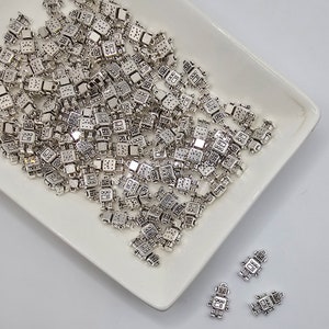May include: A white ceramic dish filled with silver robot charms. The charms are small and detailed, with a variety of robot designs. There are also three robot charms on a white surface next to the dish.