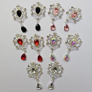 Puede incluir: Una colección de broches ornamentados con metal plateado y piedras preciosas de varios colores. Los broches presentan una piedra central rodeada de pequeños cristales y una gema colgante en forma de lágrima. Colores: negro, rosa, rojo e iridiscente.
