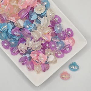 Pode incluir: Uma coleção de pingentes em forma de concha de mar iridiscente em várias cores pastel, incluindo rosa, azul e roxo. Os pingentes estão espalhados em um prato branco.