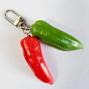 May include: A keychain featuring two pepper charms. One charm is a vibrant red pepper, and the other is a bright green pepper. The charms are attached to a silver-colored metal clasp. The peppers are smooth and glossy.