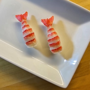 May include: Two shrimp sushi nigiri on a white rectangular plate. The shrimp is pink and white with red stripes.