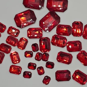 May include: A collection of red, faceted, glass gems in various sizes and shapes. The gems are scattered on a white surface.