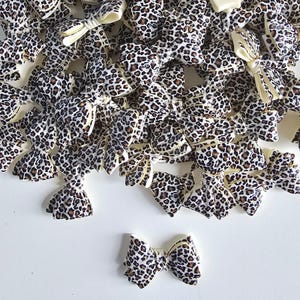 May include: A collection of small, decorative bow accents with a leopard print pattern. The bows are primarily white with brown and black spots, and some have a cream-colored center. They are scattered on a white surface.