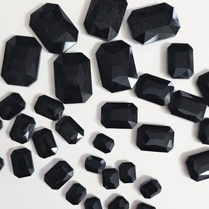 May include: A collection of black, faceted, rectangular glass gems. The gems are scattered on a white surface.