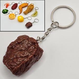 May include: A brown, textured steak keychain with a silver chain and keyring. The inset shows various food-themed keychains, including a shrimp, chicken wing, corn, and more.