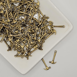 May include: A large pile of antique gold-toned hammer charms on a white ceramic plate. Two additional charms are shown separately on the plate.