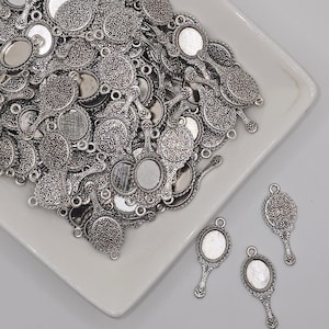 May include: A collection of silver-colored metal charms in the shape of hand mirrors and round decorative pendants. The charms have intricate floral and scrollwork patterns. Some charms have a blank oval space for customization.