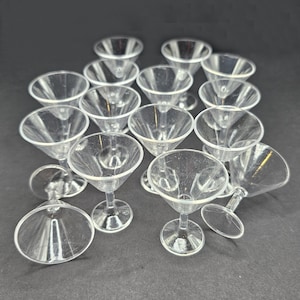 May include: A collection of clear plastic martini glasses. The glasses have a classic conical bowl shape and a stemmed base. The image shows the glasses arranged on a dark surface, with some glasses upright and others tilted.
