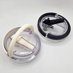 Könnte beinhalten: Zwei transparente Plastikdeckel mit einem schwarzen und weißen Silikon-Griff. Die Deckel haben einen kleinen Ausguss an der Seite und einen transparenten Plastikeinsatz in der Mitte.