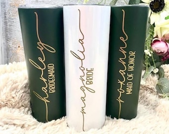 May include: Three stainless steel tumblers with gold lettering. The tumblers are green, white, and green. The text on the tumblers reads "Harley Bridesmaid", "Magnolia Bride", and "Lexanne Maid of Honor".