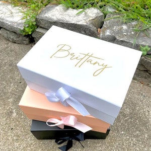 May include: A stack of three gift boxes in white, peach, and black. The white box has the name "Brittany" in gold script. Each box is tied with a satin ribbon in white, pink, and black. The boxes are stacked on a concrete surface.