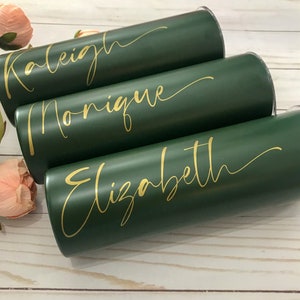 May include: Three green tumblers with gold lettering. The names "Raleigh", "Monique", and "Elizabeth" are written on the tumblers.