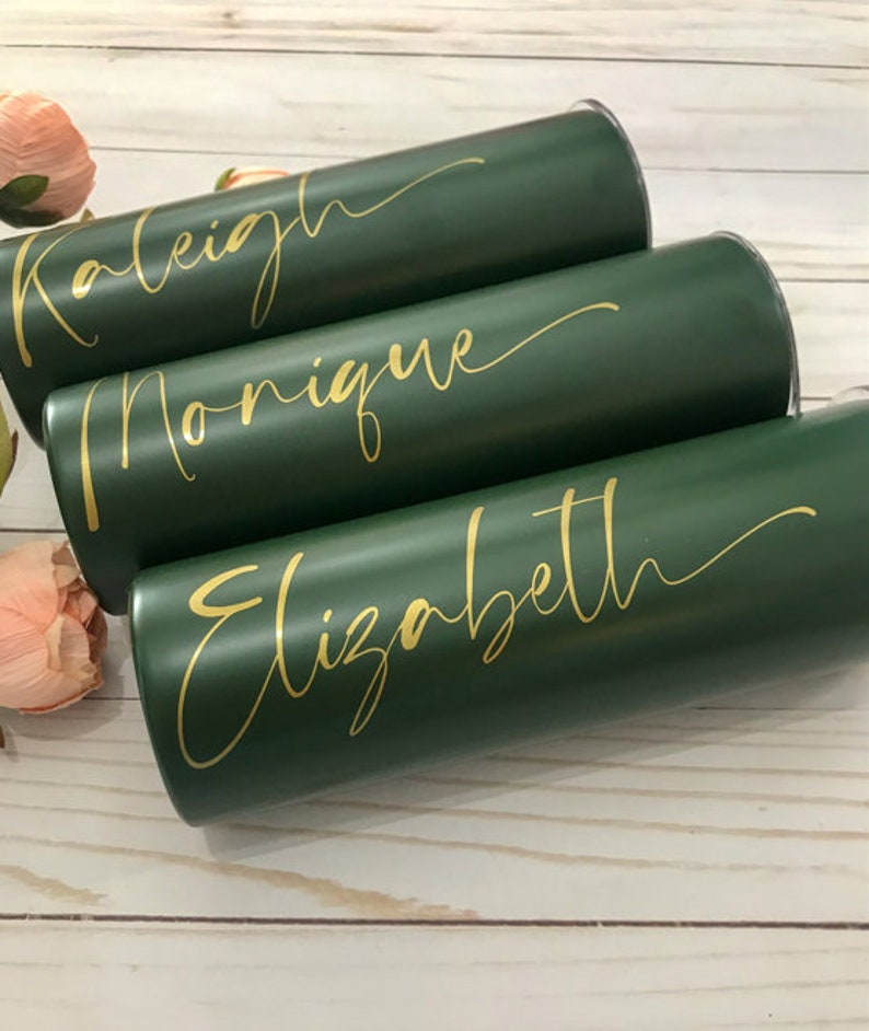 May include: Three green tumblers with gold lettering. The names "Raleigh", "Monique", and "Elizabeth" are written on the tumblers.