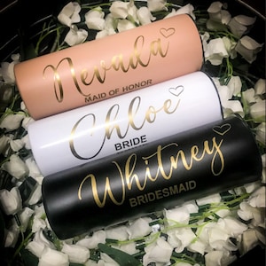 May include: Three stainless steel tumblers with gold lettering. The top tumbler is peach coloured with the name "Nevada" and the title "Maid of Honour". The middle tumbler is white with the name "Chloe" and the title "Bride". The bottom tumbler is black with the name "Whitney" and the title "Bridesmaid".
