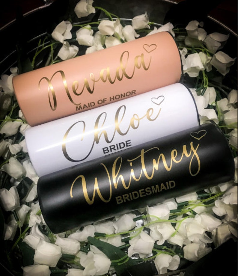 May include: Three stainless steel tumblers with gold lettering. The top tumbler is peach coloured with the name "Nevada" and the title "Maid of Honour". The middle tumbler is white with the name "Chloe" and the title "Bride". The bottom tumbler is black with the name "Whitney" and the title "Bridesmaid".