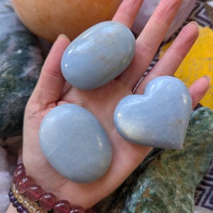 Pode incluir: Três pedras de celestita azuis, uma em forma de coração e duas ovais, em uma mão.
