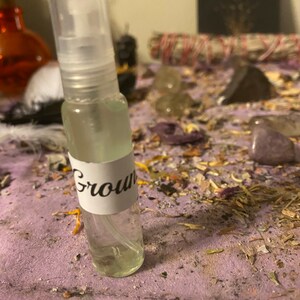 May include: A clear glass bottle with a white label that says "Ground" on it. The bottle has a spray nozzle and is sitting on a purple surface with various dried herbs and crystals.