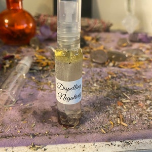 May include: A clear glass bottle with a spray nozzle and a white label that reads "Dispelling Negativity". The bottle is filled with a clear liquid and is sitting on a purple surface.