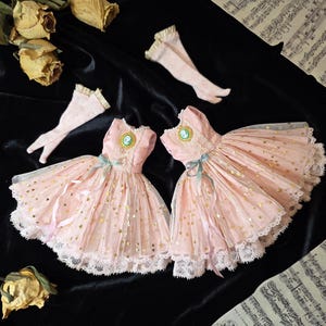 Pode incluir: Dois vestidos sem mangas em rosa pálido com sobreposição de tule com estrelas douradas, bainha de renda, um laço central na cintura e um broche de camafeu no decote, acompanhados por dois pares de meias a condizer com acabamento em renda.