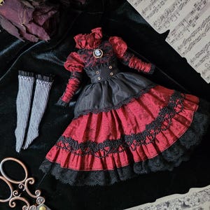 May include: A miniature red and black dress with lace trim, a high collar, and gold buttons. The dress is accompanied by striped socks and a pair of vintage scissors. A dried rose and sheet music are also in the image.