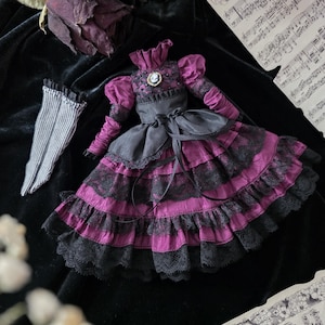 May include: A doll-sized, Victorian-style dress in shades of purple and black, featuring lace detailing and a ribbon sash. The dress has long sleeves and a high collar, with a cameo brooch. A pair of striped gloves and sheet music are also visible.