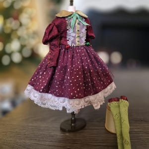Può includere: Un vestito da bambola bordeaux con finiture in pizzo bianco e colletto dorato, esposto su un manichino in miniatura. L'abito presenta un pannello in pizzo bianco ed è abbinato a calze verde chiaro e un cappello piccolo.