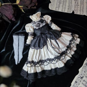 May include: A miniature black and ivory dress with lace detailing, a high collar, and puff sleeves. The doll dress has a black bodice and a cream-colored skirt with black lace trim. A pair of gray and white striped doll stockings are also present.