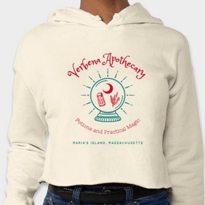 May include: A cream-colored cropped hoodie with a graphic design of a teal crystal ball with a crescent moon, a sprig of herbs, and a salt shaker inside. The text "Verbena Apothecary" is in red and teal, and the text "Potions and Practical Magic" is in teal. The text "Maria's Island, Massachusetts" is in black.