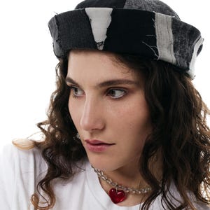 May include: A woman with curly brown hair is wearing a black and white patchwork hat and a white t-shirt with a red heart necklace.