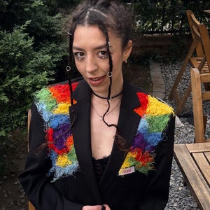 Puede incluir: Una chaqueta negra con un parche de crochet de colores arcoíris en la parte delantera. El parche está hecho de hilo y tiene un borde deshilachado. La chaqueta tiene una pequeña etiqueta que dice "#ssbsly".