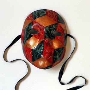 May include: A decorative mask with a harlequin pattern in red, green, and gold. The mask has eye holes and a nose indentation. Black ribbons are attached on each side for securing the mask.