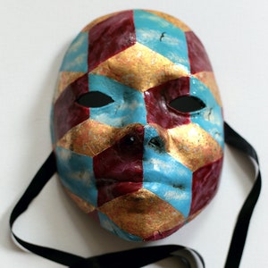 Könnte beinhalten: Eine farbenfrohe Maske mit einem geometrischen Muster aus roten, blauen und goldenen Formen. Die Maske hat Augenlöcher und eine Nase, mit einem schwarzen Band zum Tragen. Die Maske ist wahrscheinlich für Kostüm- oder Dekorationszwecke gedacht.
