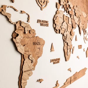 Pode incluir: Um mapa-m&uacute;ndi de madeira detalhado com contornos de pa&iacute;ses e nomes de oceanos. O mapa inclui uma rosa dos ventos e pequenos detalhes de veleiros. Os continentes s&atilde;o em um tom marrom quente, montados em uma parede branca.