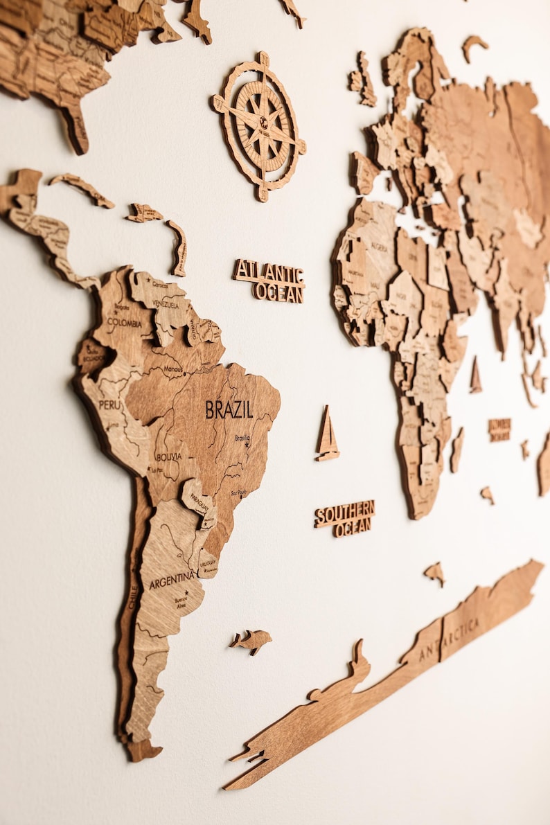 Pode incluir: Um mapa-m&uacute;ndi de madeira detalhado com contornos de pa&iacute;ses e nomes de oceanos. O mapa inclui uma rosa dos ventos e pequenos detalhes de veleiros. Os continentes s&atilde;o em um tom marrom quente, montados em uma parede branca.
