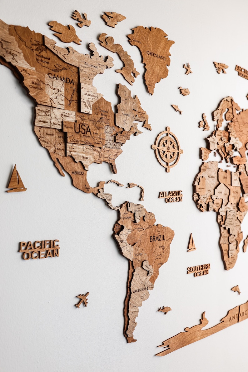 Pode incluir: Um mapa-m&uacute;ndi de madeira com contornos detalhados de pa&iacute;ses e r&oacute;tulos de oceanos. O mapa apresenta uma rosa dos ventos e pequenos detalhes de madeira, como avi&otilde;es e veleiros. O mapa &eacute; feito de madeira marrom clara.