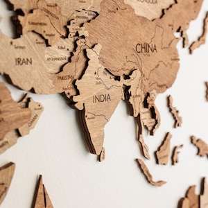 Pode incluir: Um mapa-m&uacute;ndi de madeira detalhado, cortado a laser de madeira castanha clara, montado numa parede branca. O mapa destaca a &Aacute;sia, com pa&iacute;ses como &Iacute;ndia, China e Ir&atilde;o claramente rotulados. Inclui um veleiro de madeira e as palavras "INDIAN OCEAN".