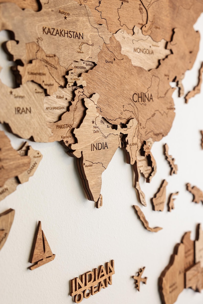 Pode incluir: Um mapa-m&uacute;ndi de madeira detalhado, cortado a laser de madeira castanha clara, montado numa parede branca. O mapa destaca a &Aacute;sia, com pa&iacute;ses como &Iacute;ndia, China e Ir&atilde;o claramente rotulados. Inclui um veleiro de madeira e as palavras "INDIAN OCEAN".
