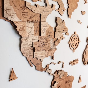 Pode incluir: Um mapa-m&uacute;ndi de madeira com contornos detalhados de pa&iacute;ses, incluindo EUA, Canad&aacute; e M&eacute;xico. O mapa &eacute; feito de madeira castanha clara e inclui pe&ccedil;as separadas para continentes e ilhas. Elementos decorativos como uma b&uacute;ssola e veleiros tamb&eacute;m est&atilde;o presentes.