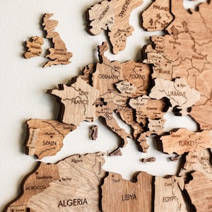 May include: A wooden world map showing Europe and Africa, with country names engraved. The map is made of light-coloured wood, with detailed borders and city names. The United Kingdom, France, and Spain are clearly visible.