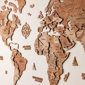 Pode incluir: Um mapa-m&uacute;ndi de madeira detalhado, com continentes e pa&iacute;ses, montado numa parede branca. Inclui elementos decorativos como uma rosa dos ventos, veleiros e criaturas marinhas. O mapa &eacute; feito de madeira castanha clara.