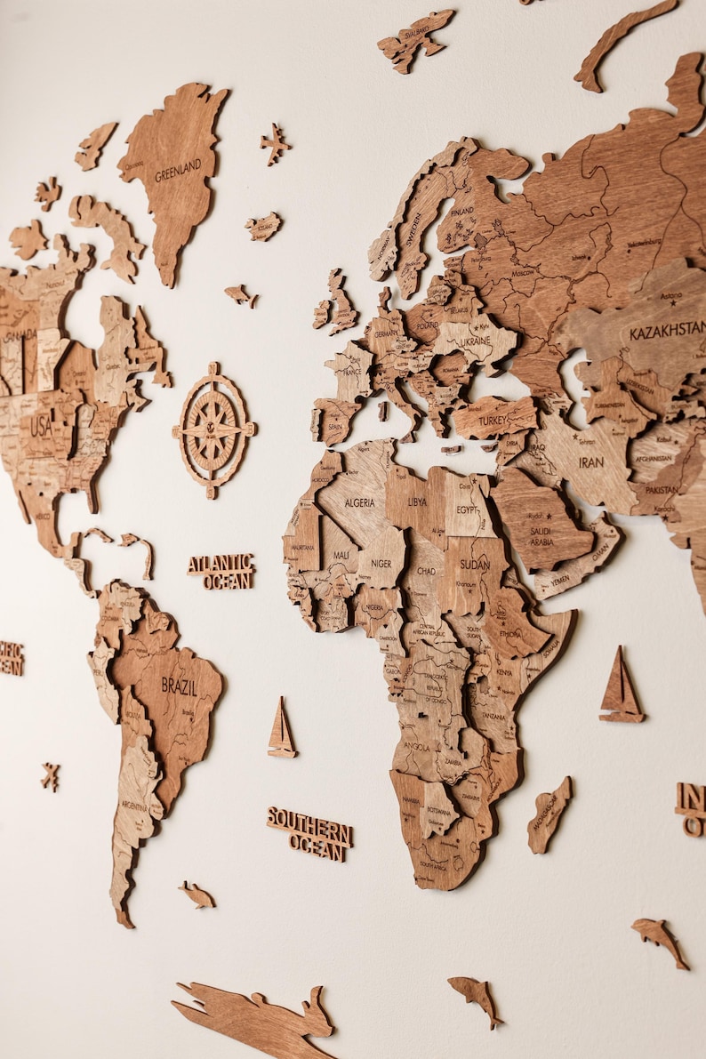Pode incluir: Um mapa-m&uacute;ndi de madeira detalhado, com continentes e pa&iacute;ses, montado numa parede branca. Inclui elementos decorativos como uma rosa dos ventos, veleiros e criaturas marinhas. O mapa &eacute; feito de madeira castanha clara.