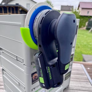 Könnte beinhalten: Ein grün-schwarzer Festool ETC 125 18V Schleifer mit einem grünen Akku. Der Schleifer hängt an einem grauen Aufbewahrungsbehälter mit einer grün-blauen Schleifscheibe.