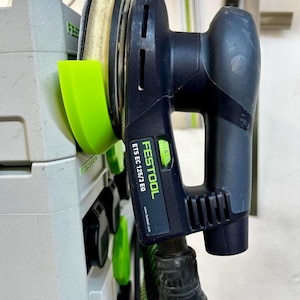 Könnte beinhalten: Ein schwarzer und grauer Festool ET5 EC 125/3 EQ-Schwingschleifer mit grünem Einschaltknopf und beigem Schleifpad. Der Schleifer ist an einem grünen und schwarzen Schlauch befestigt und an einem grauen Aufbewahrungsbehälter montiert.