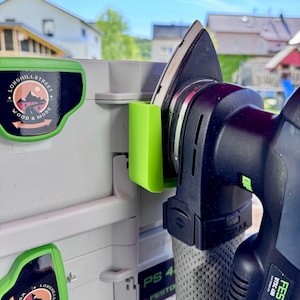 Könnte beinhalten: Nahaufnahme eines schwarzen und grauen Elektroschleifers mit einem grünen Halter, der an einem grauen Aufbewahrungsbehälter befestigt ist. Der Schleifer zeigt sein Schleifpad und seinen Griff. Der Behälter hat einen Aufkleber mit der Aufschrift "Longhill Street Wood & More".